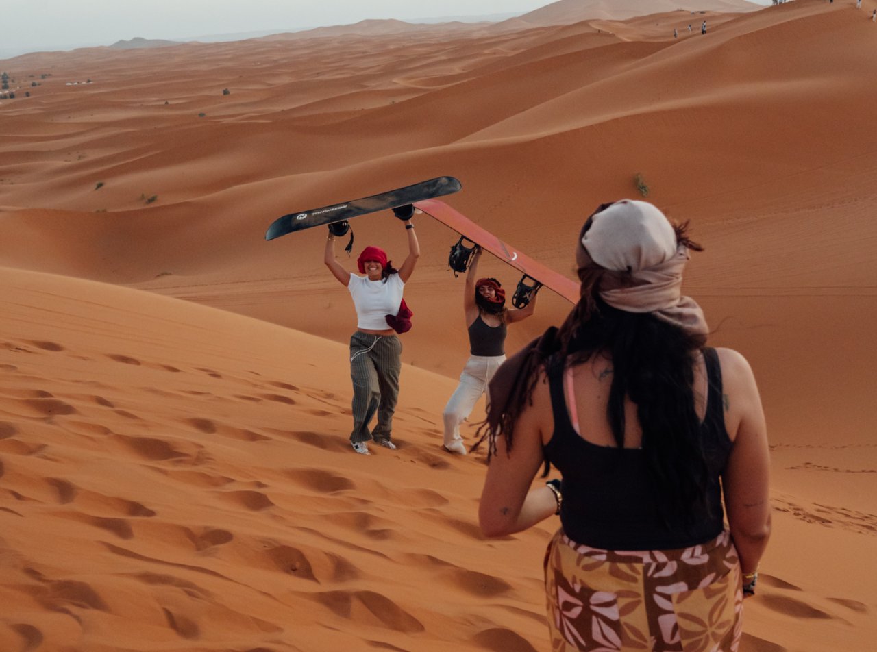 Three young travellers carrying sandboards hike up a dune in the Sahara Desert, smiling and wrapped in desert scarves, surrounded by sand dunes under the evening sky in Morocco.