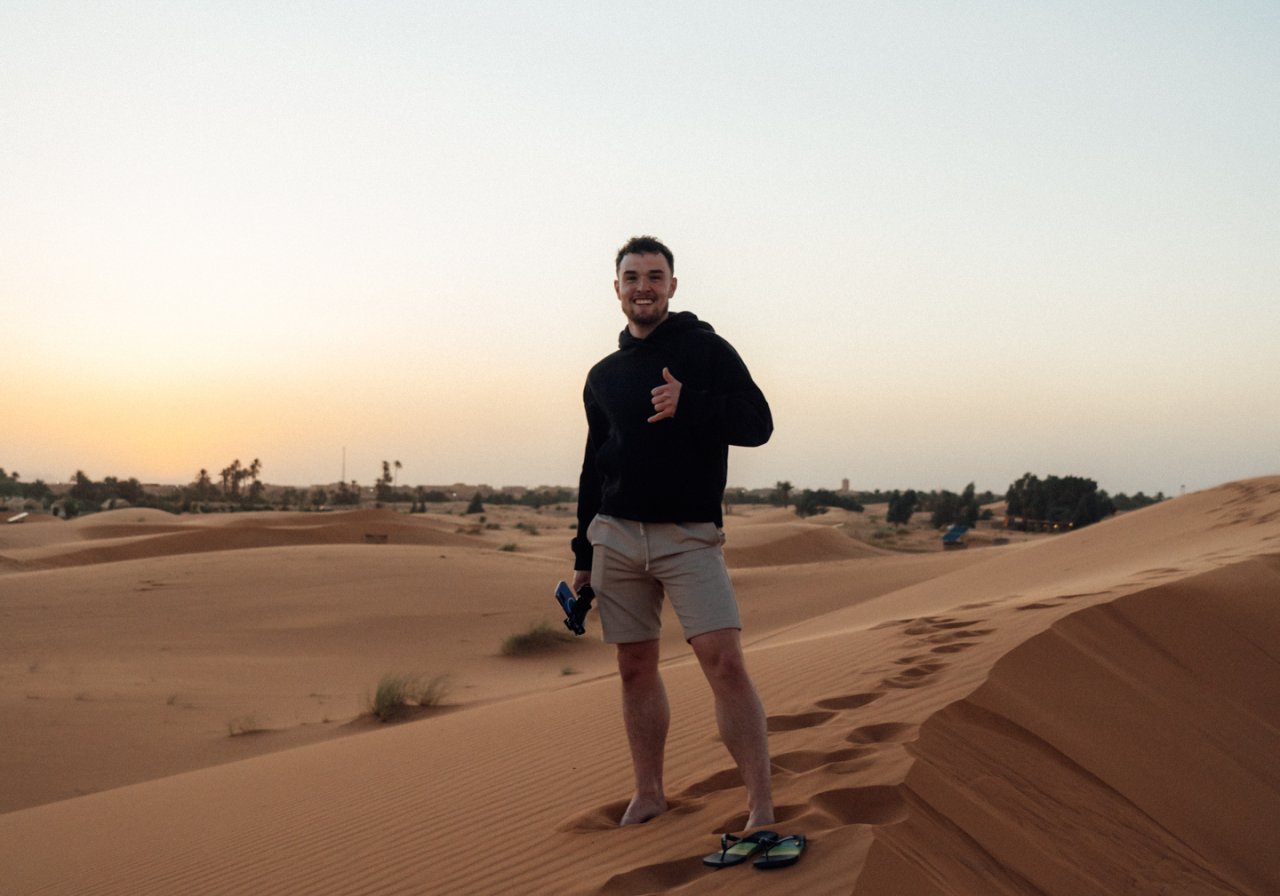 Young male traveller poses in the dunes of the Sahara desert with a camera in hand ready to film content